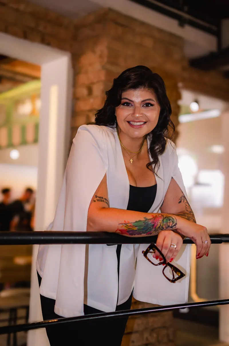 Kelly Pinheiro de blazer branco, apoiada em um guarda-corpo, segurando os óculos e sorrindo