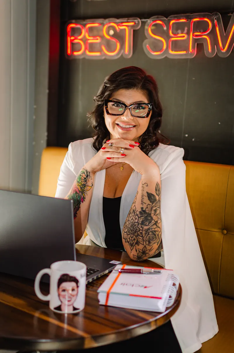 Kelly Pinheiro de blazer branco, sorrindo com as mãos sob o queixo, diante de um notebook e uma caneca com a própria caricatura, sob um letreiro neon