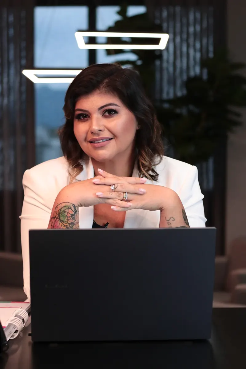 Kelly Pinheiro de blazer branco, mãos cruzadas sobre o notebook, em uma sala de fundo escuro