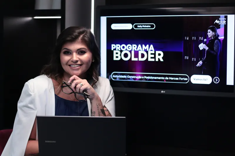 Kelly Pinheiro segurando os óculos, sorrindo, com uma TV ao fundo exibindo o Programa Bolder